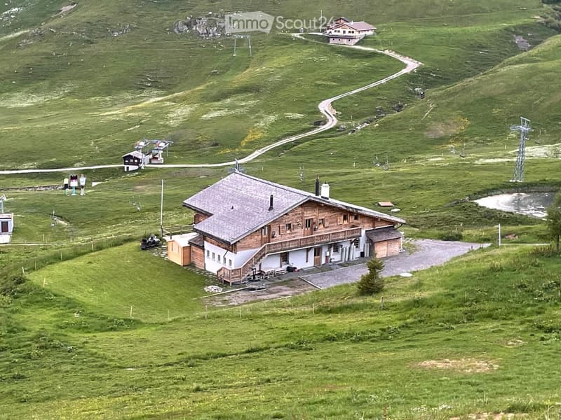 Mountain house, wooden exterior, gray roof, surrounded by green hills, driveway, small pond
