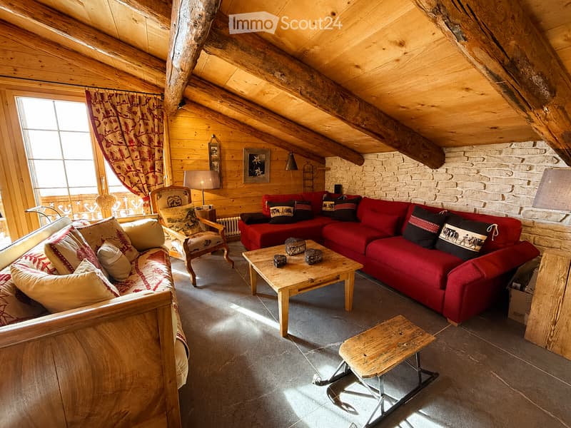 Cozy living room with wooden beams, exposed stone wall, large window with red curtains, red sectional sofa, wooden coffee table, armchair, and small wooden stool.