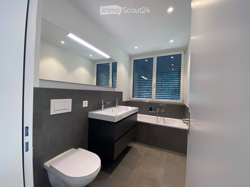 modern bathroom with bathtub, sink, toilet, large mirror, gray tiles, white ceiling, window with shutters