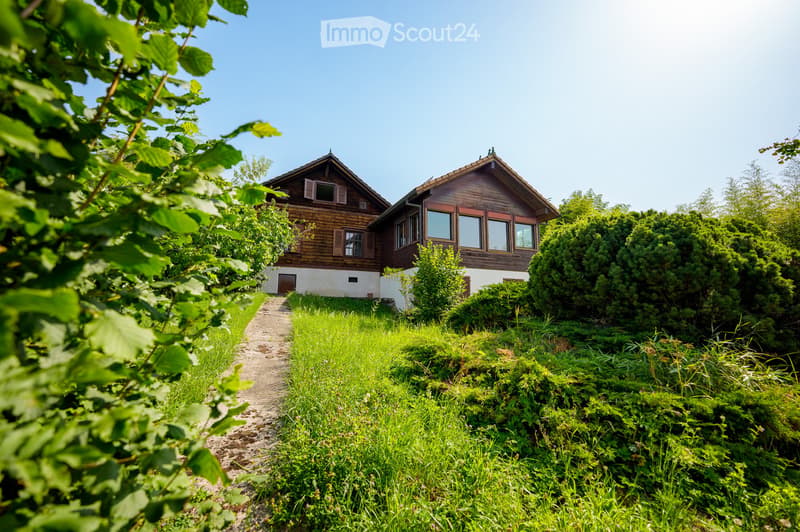 Two-story wooden house, surrounded by greenery, driveway leading to the entrance, situated in a rural area.