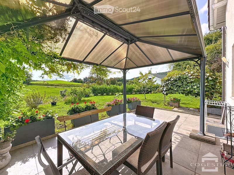 Outdoor dining area with a glass table and chairs, covered by a pergola, surrounded by green grass and plants, with a view of a green field.