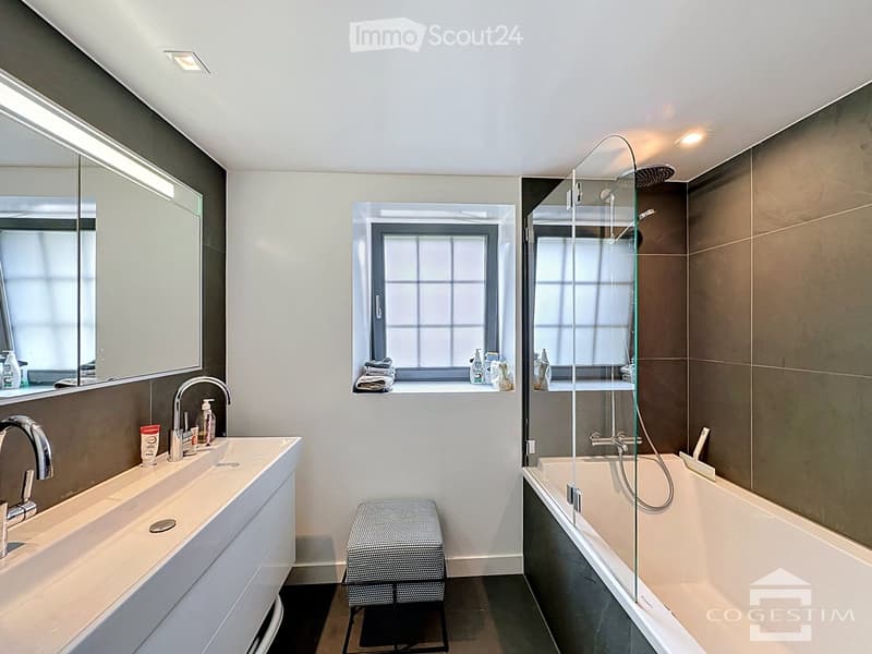 Modern bathroom with a double sink, bathtub with glass shower enclosure, and a window with towels and toiletries on the sill.