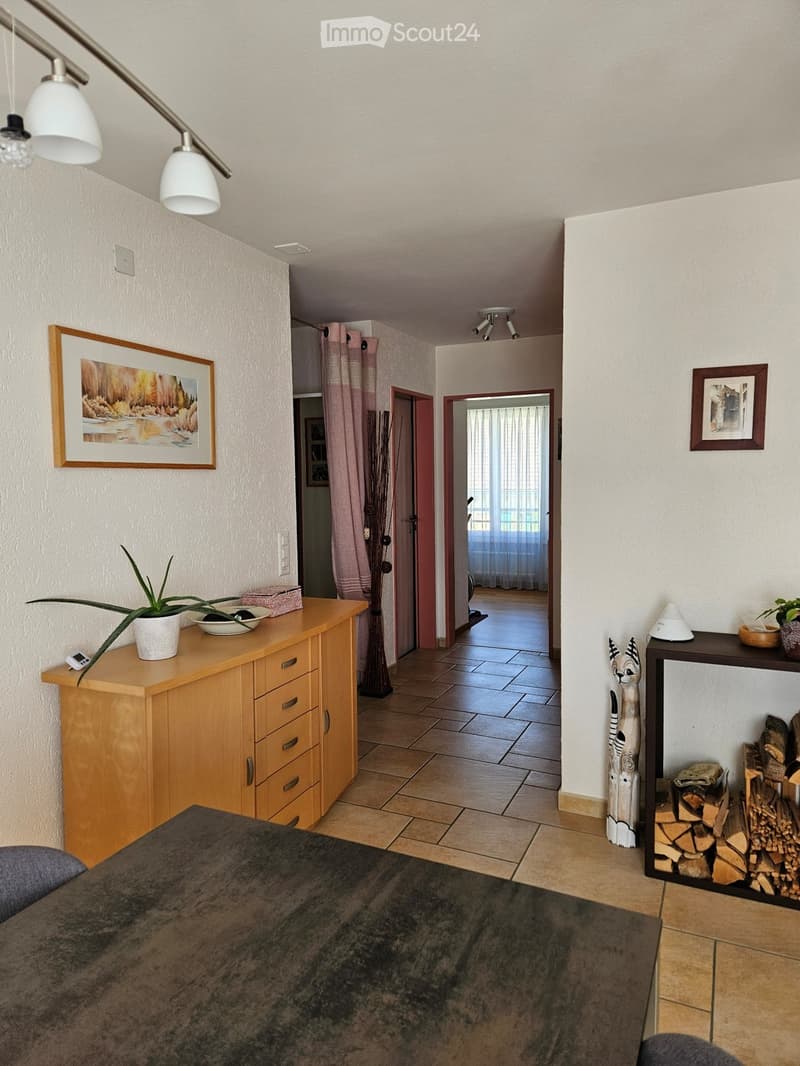 room has wooden floor, wooden sideboard, white walls, mounted frames, wooden table, potted plants, lamp, fireplace, window