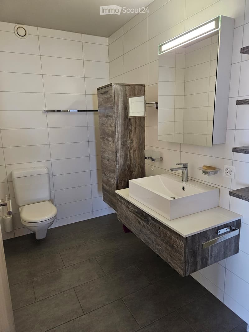 Bathroom with white walls, black tiled floor, wooden cabinet, sink, faucet, mirror with lighted frame, toilet, shelves, and electrical outlet.