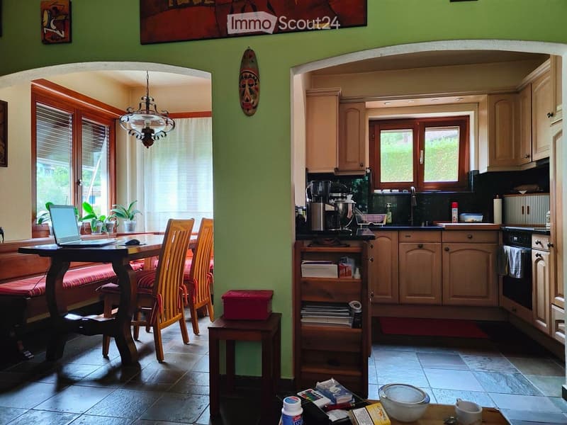 Dining area with a wooden table, chairs, and a laptop. Kitchen with wooden cabinets, a sink, and a coffee machine. A wooden shelf with books and a red box. A green wall with a mask and a painting.