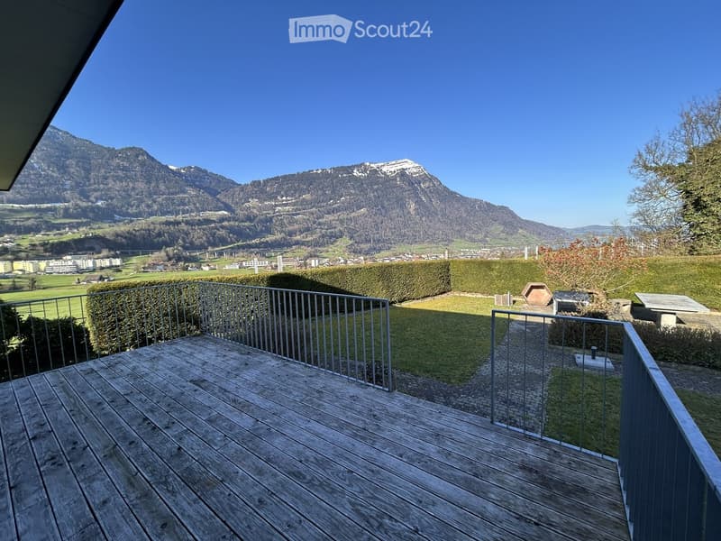 wooden deck, metal railing, green grass, mountains, clear sky