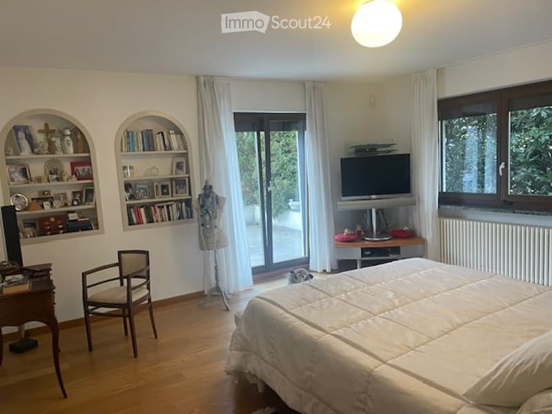 large bed, wooden floor, white walls, large windows, sliding glass doors, shelves with books and items, chair, desk, television, radiator