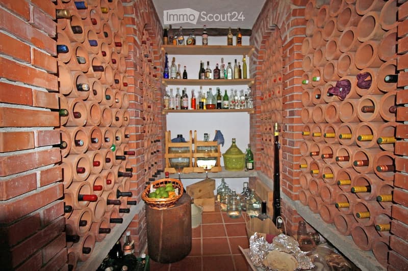 Wine cellar with brick walls, shelves with bottles, wooden barrel, baskets with fruits, glass jars, and a wooden box.