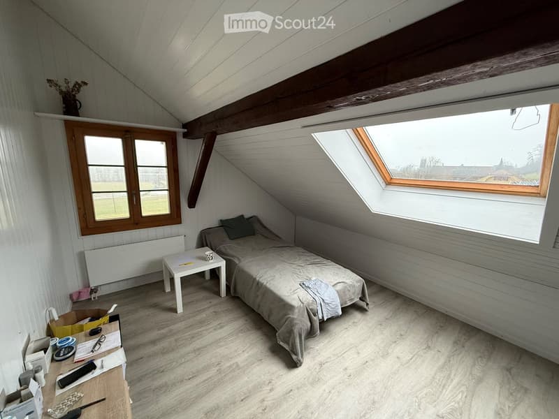 A small bedroom with a single bed, desk, small window, slanted ceiling and attic window.