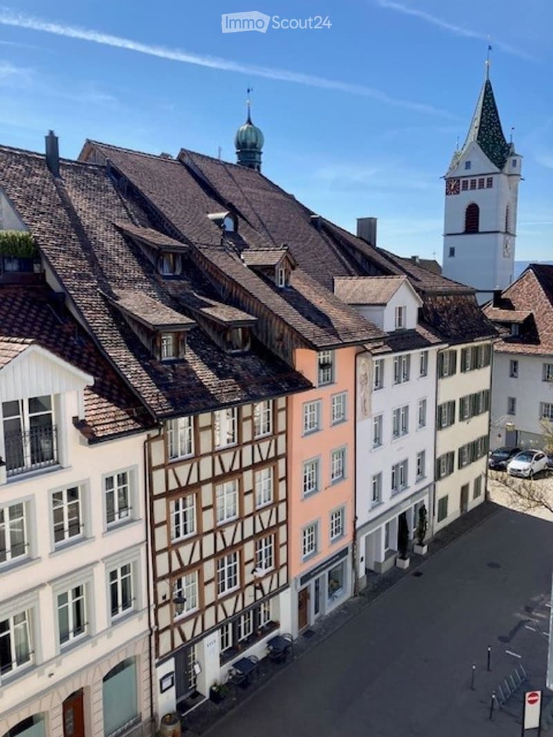 A row of buildings with brown roofs, white walls, and many windows. The buildings are of different sizes and styles, with some having balconies and others having chimneys. The street is empty, and there are cars parked on the side. In the distance, there