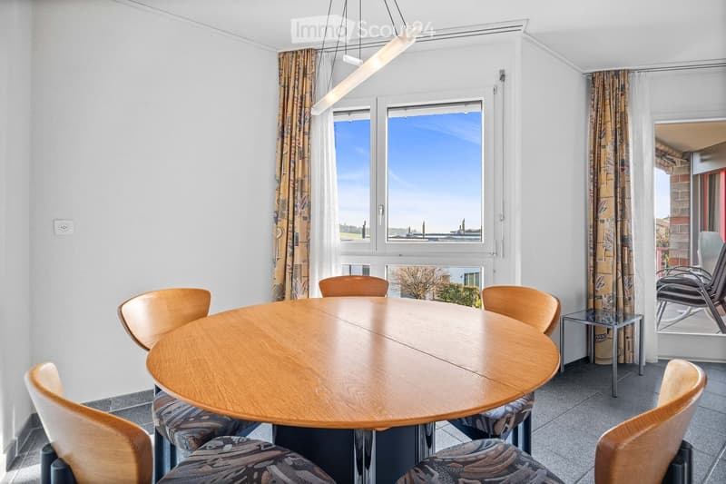 Dining area with a round wooden table, 6 chairs, large windows, curtains, and a view of the outside