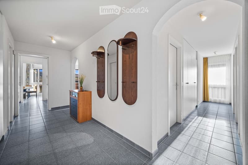 Empty hallway with white walls, gray tiles, wooden cabinet, three wall-mounted coat racks, and a mirror.