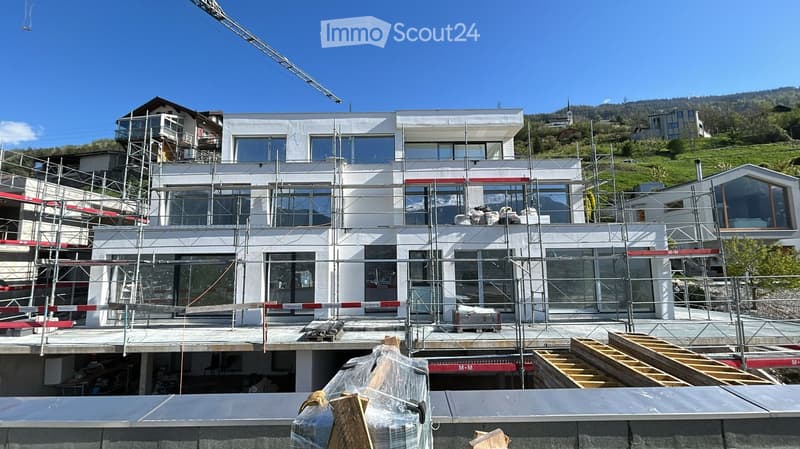 Modern house under construction, white exterior, large windows, scaffolding, crane, surrounded by other buildings and green hills