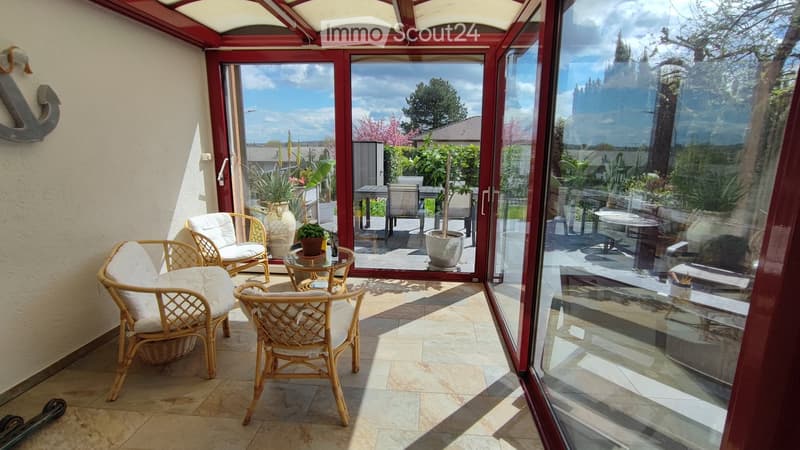Four rattan chairs, glass table with green plants, two vases with plants, two large glass doors