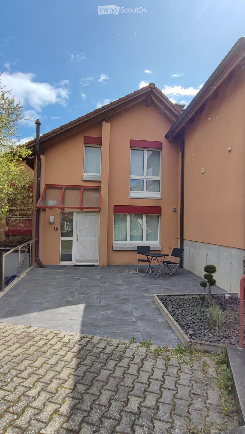 2 story house, brown exterior, white door, red trim, windows, patio with table and chairs, brick walkway, small garden