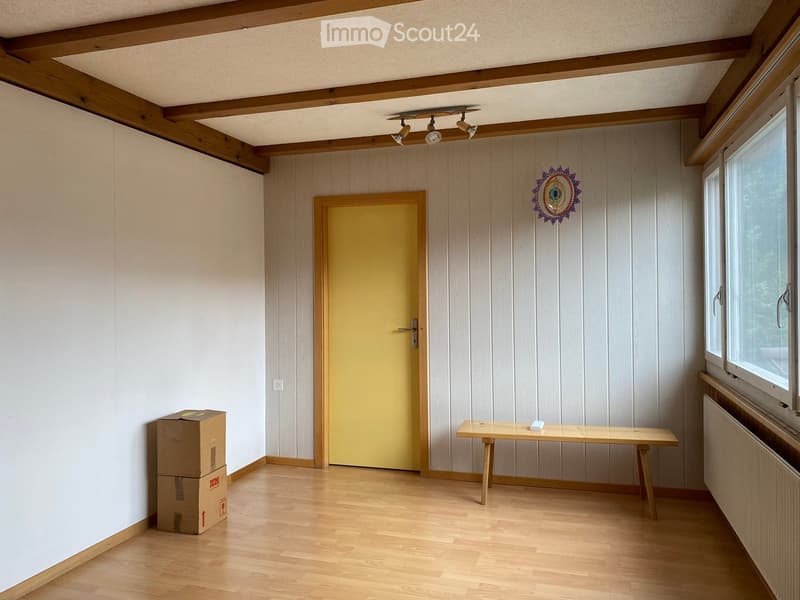 Empty room with wooden floor, white walls, yellow door, wooden bench, ceiling lights, and a window with a view of trees outside.