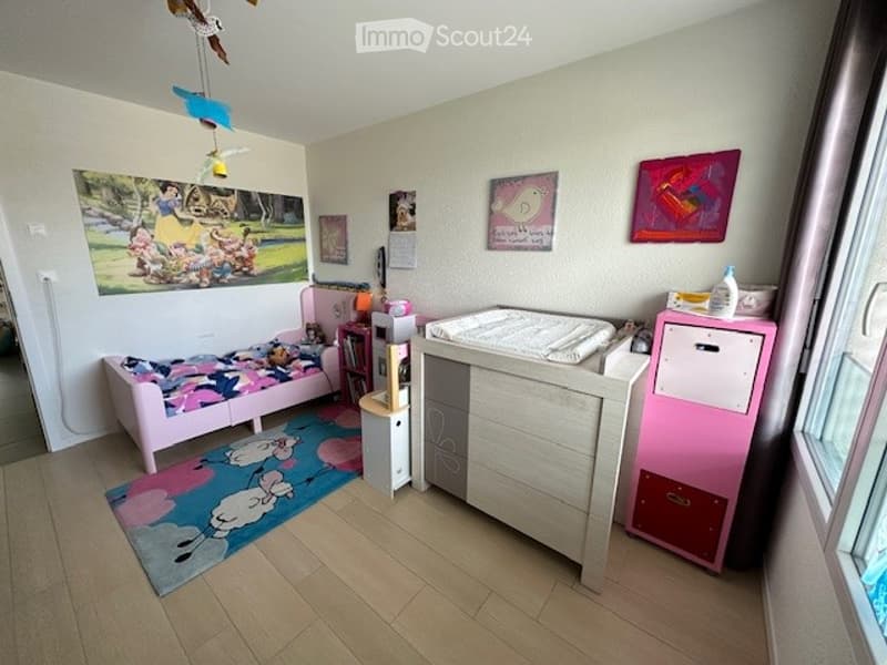 A child's bedroom with a pink bed, a crib, a rug, a pink cabinet, a bookshelf, and a wall with paintings.