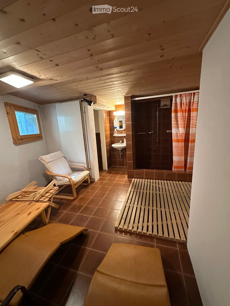 Small bathroom with wooden bench, chair, and table. Brown tiled floor and walls. Shower with curtain, sink, and mirror.