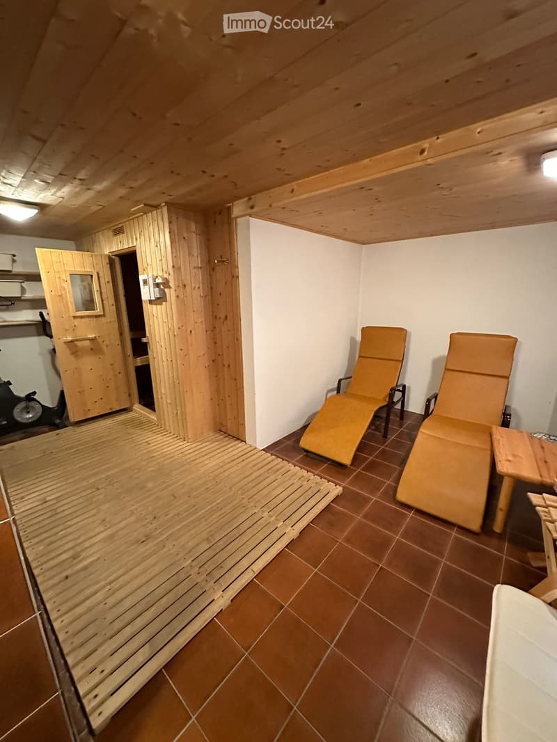 A room with a wooden sauna, exercise bike, two lounge chairs, a wooden table, and a wooden bench.