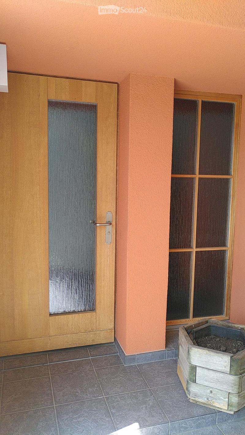 A narrow hallway with peach-colored walls, a wooden door with a frosted glass panel, a window with wooden frame, and a small wooden planter on the floor.