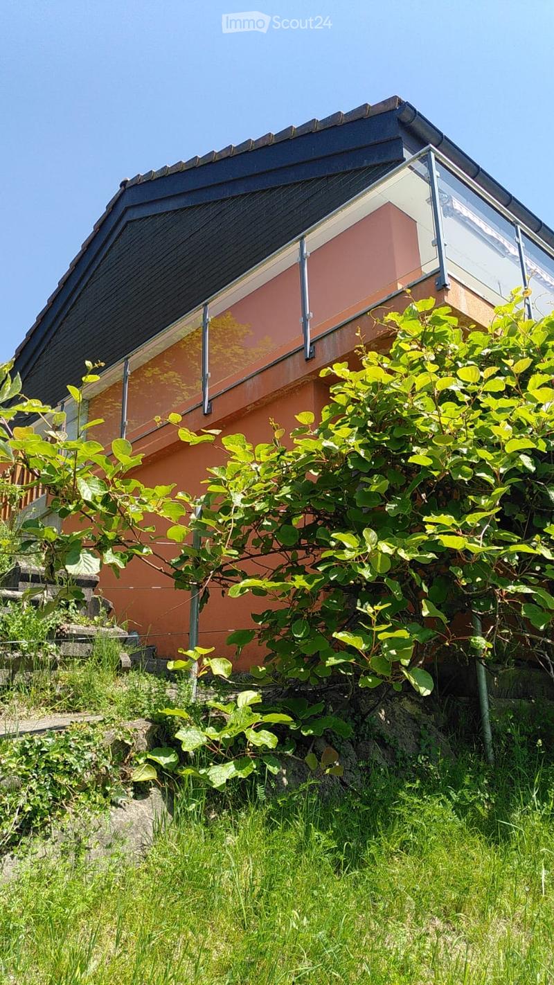 A two-story house with a modern design, featuring a sloped roof, glass balcony, and lush greenery in the foreground.