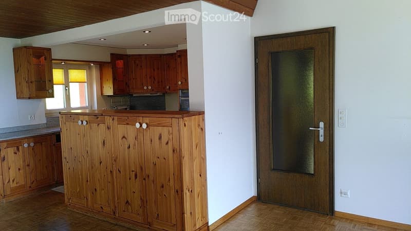 The image shows a kitchen with wooden cabinets, a countertop, and a door leading to another room. The kitchen features a wooden floor and white walls.