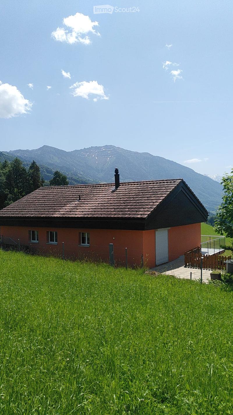 A single-story house with a red-tiled roof and orange walls, surrounded by a grassy field and mountains in the background.