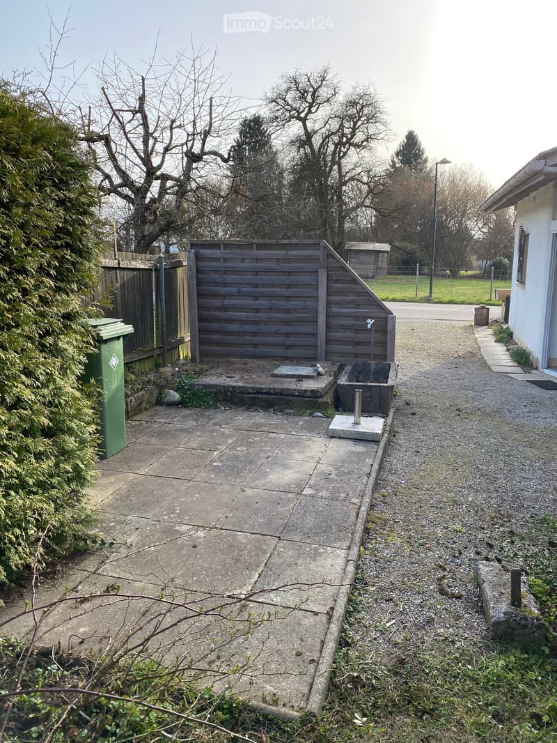 A backyard with a wooden fence, a concrete pathway, a green trash bin, a wooden structure, and a small concrete platform with a water tap.
