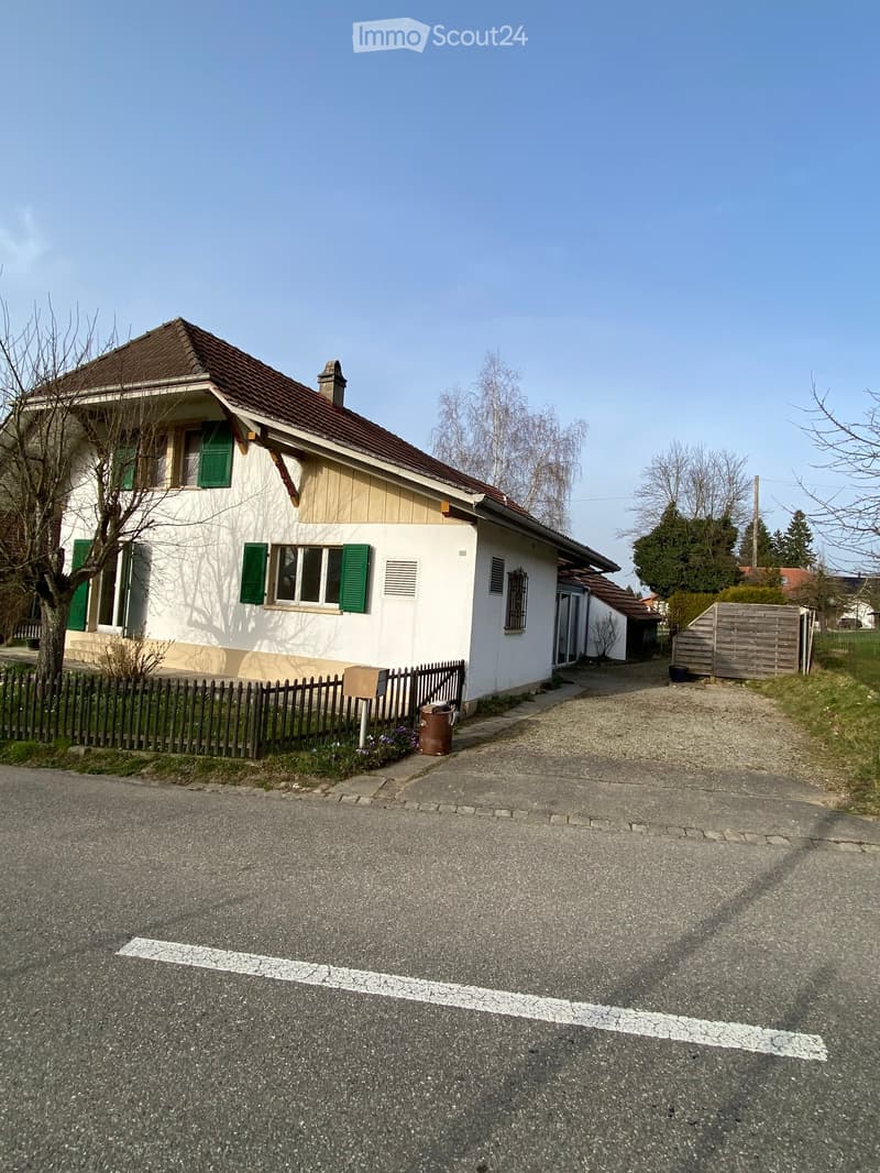 Single-family house, white exterior, brown roof, green shutters, driveway, wooden fence, mailbox, trash bin, small garden, trees, clear sky