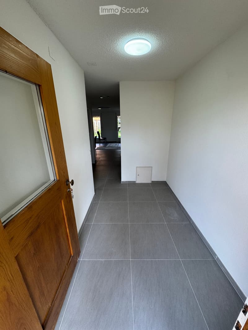 Empty hallway with wooden door, white walls, gray tiles, ceiling light