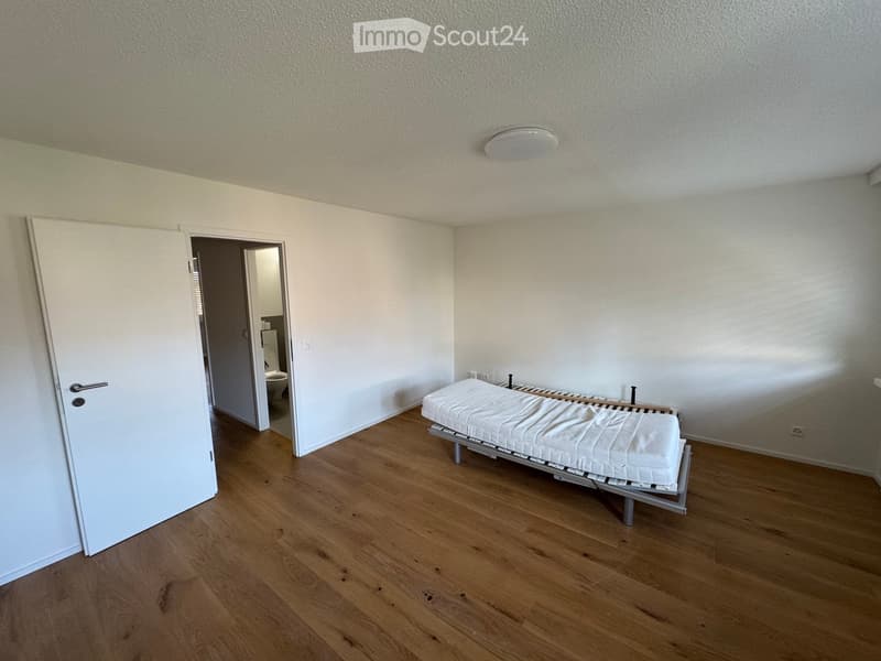 Empty room with white walls, wooden floor, white door, single bed, and a small bathroom with a toilet