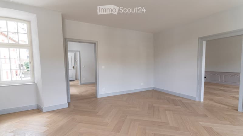 Empty room with white walls, wooden parquet floor, large window, two doors leading to other rooms