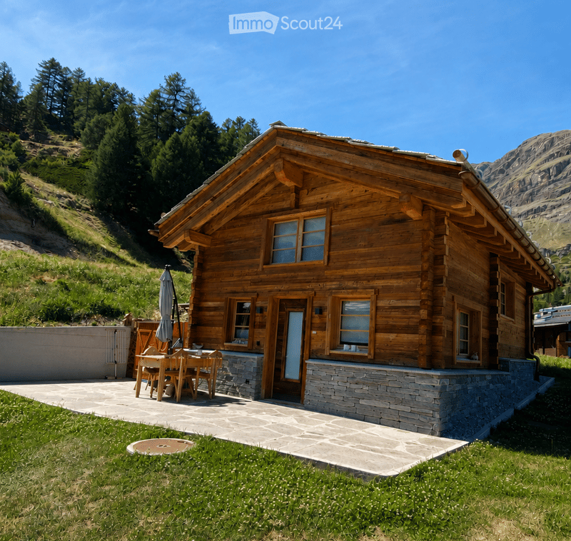 A wooden chalet-style house with a stone base, large windows, and a raised ground floor. The house is surrounded by a grassy area and a stone patio with outdoor furniture.
