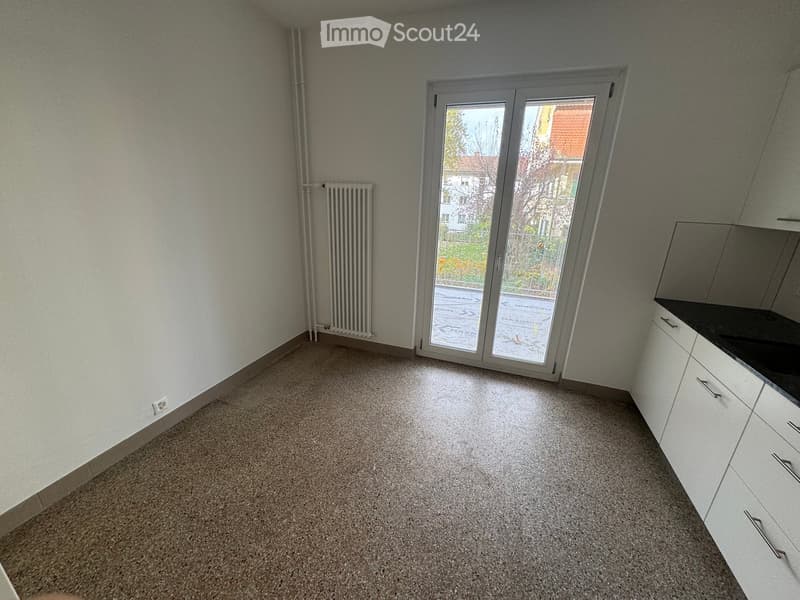 Empty room, grey floor, white walls, large sliding glass door, radiator, electrical outlet, kitchen area with white cabinets and black countertops