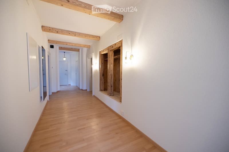 Empty hallway with wooden floor, white walls, wooden beams on ceiling, wall-mounted lights, and a wooden closet.
