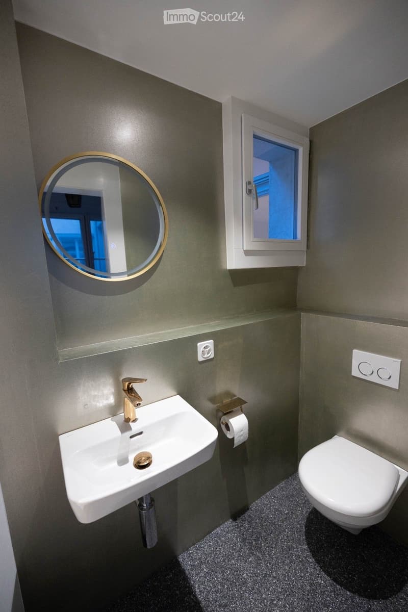 small bathroom with white sink, gold faucet, white toilet, round mirror, small window, gray walls