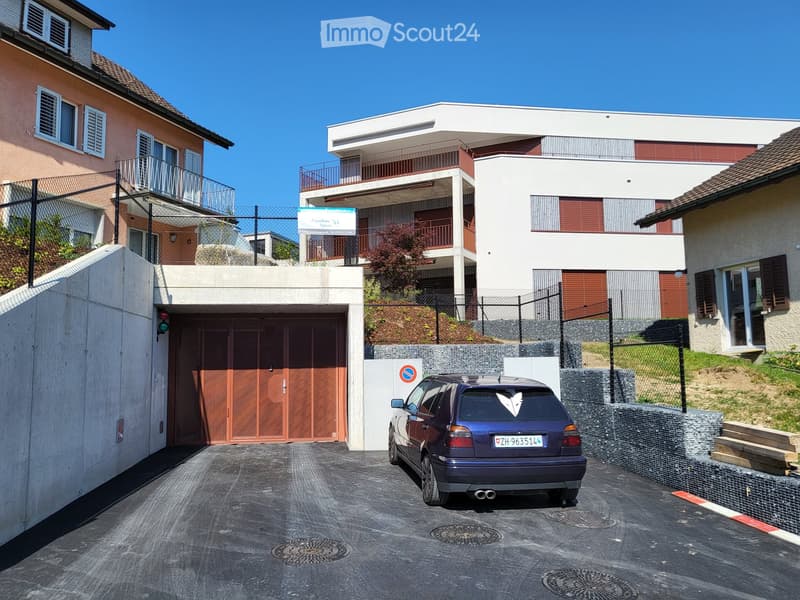 A modern apartment building with a car parked in the garage, concrete walls, and a sloped driveway.