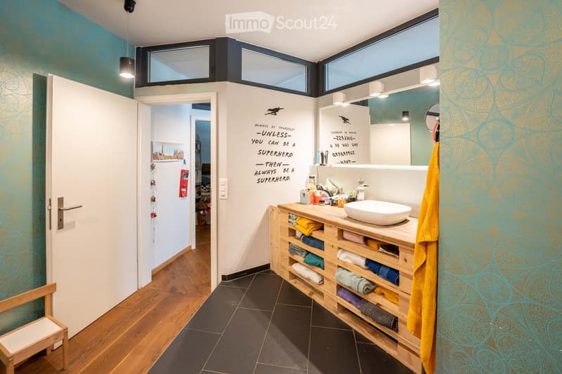 Bathroom with wooden cabinet, white sink, blue walls, wooden floor, white door, wooden chair, mirror, lamps, towel, and motivational quote.