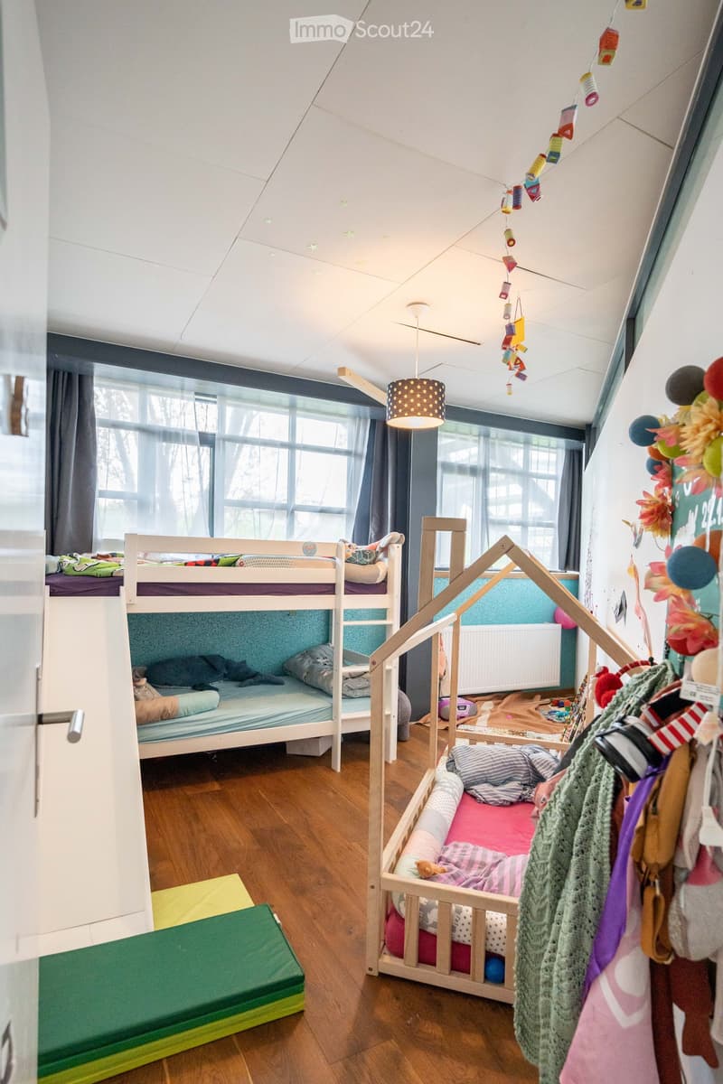 A children's room with wooden floor, bunk beds, a small wooden crib, a hanging lamp, a radiator, and a wall with decorations.