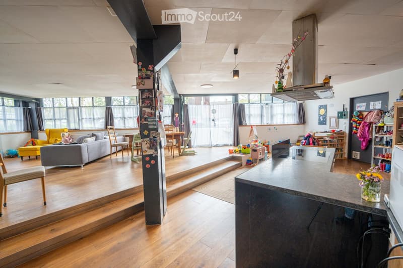 Open plan living and dining area with wooden floors, large windows, a gray sofa, a yellow armchair, a dining table, and a kitchen island with a flower vase.