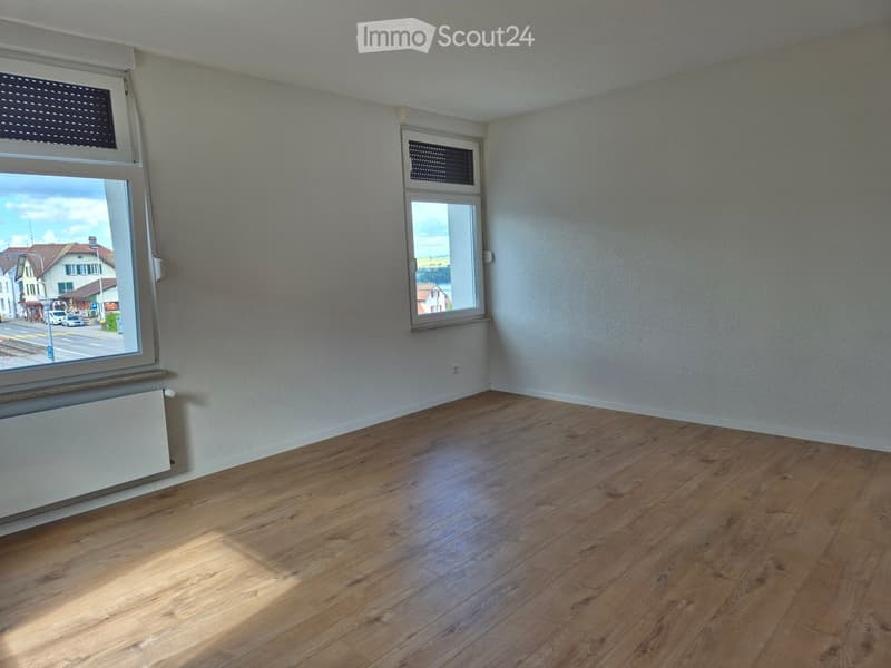 Empty room with wooden flooring, white walls, and two windows offering a view of the street and surrounding buildings.