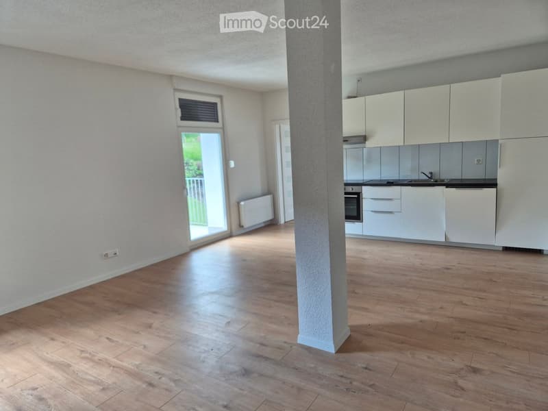 Empty room with wooden flooring, white walls, and a central pillar. A kitchen area is visible in the background with white cabinets and appliances.