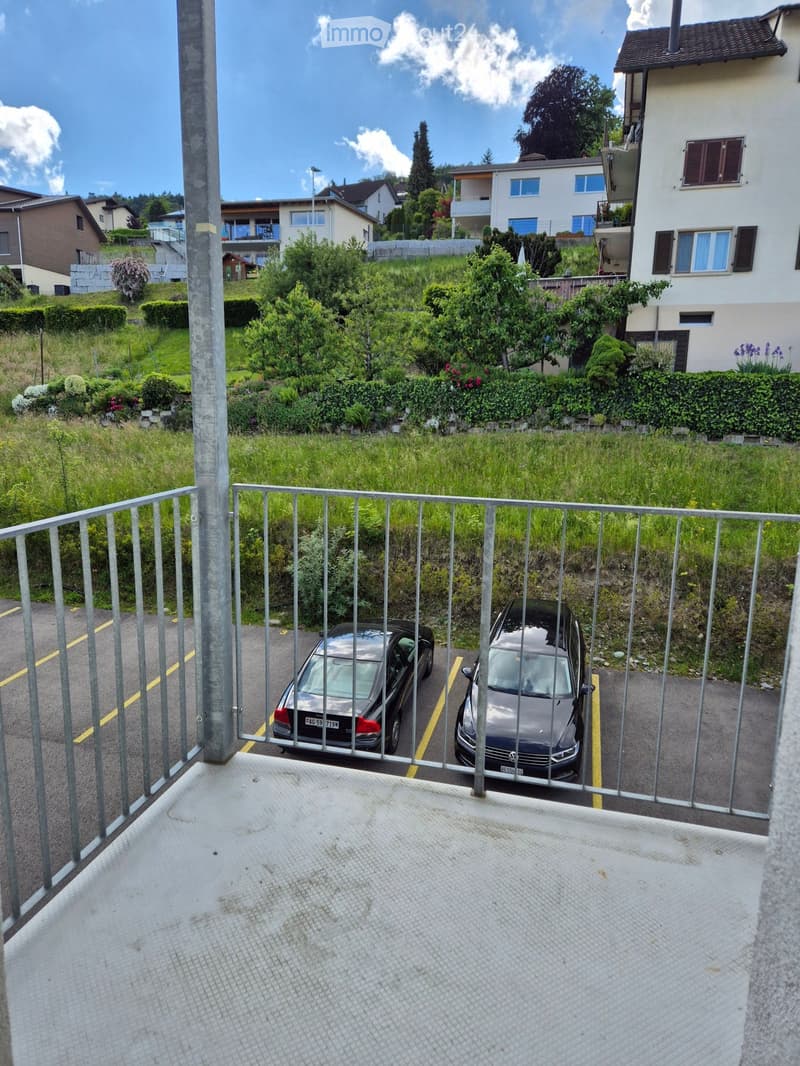 Balcony overlooking a parking area with two parked cars, surrounded by greenery and neighboring buildings.