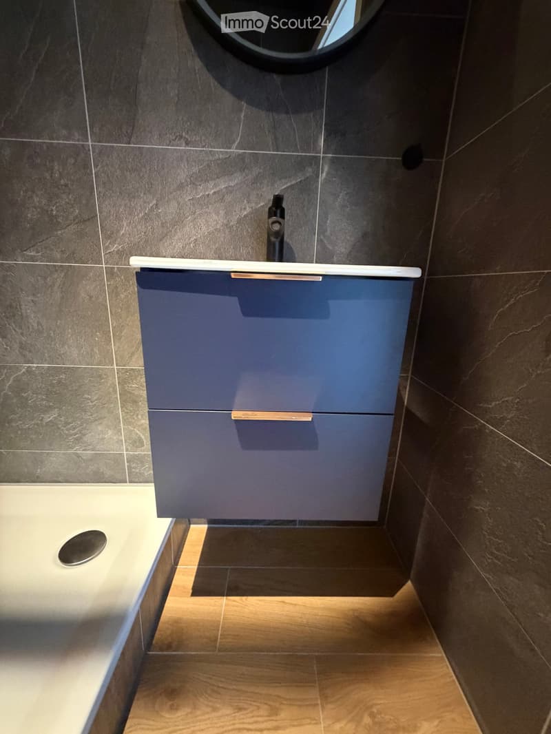 Bathroom with blue cabinet, white countertop, black faucet, tiled walls, wooden floor, and a shower area.