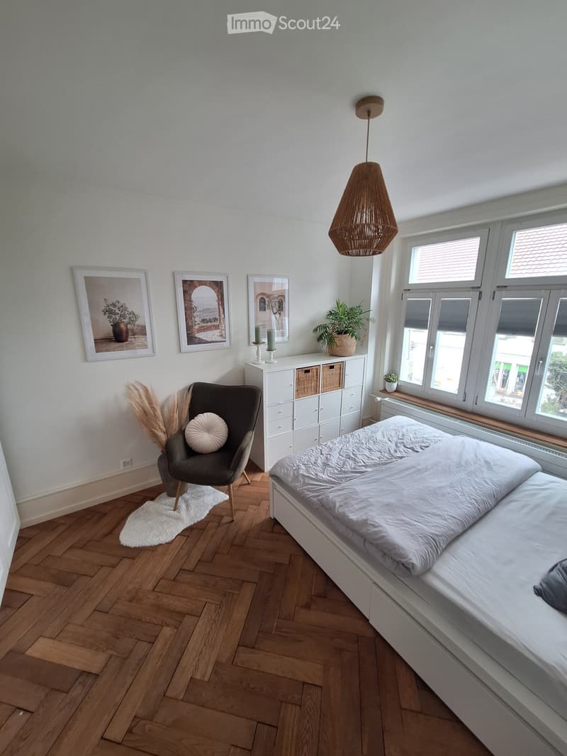 Bedroom with wooden parquet floor, white walls, bed, chair, cabinet, plants, and pictures on the wall.