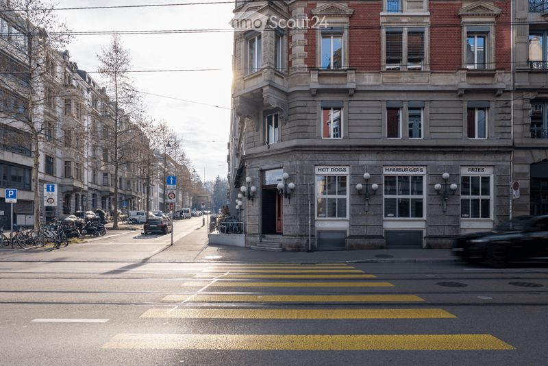 Corner building with restaurant on the ground floor, brick facade, large windows, street lamps, parked cars, bicycles, and a pedestrian crossing.
