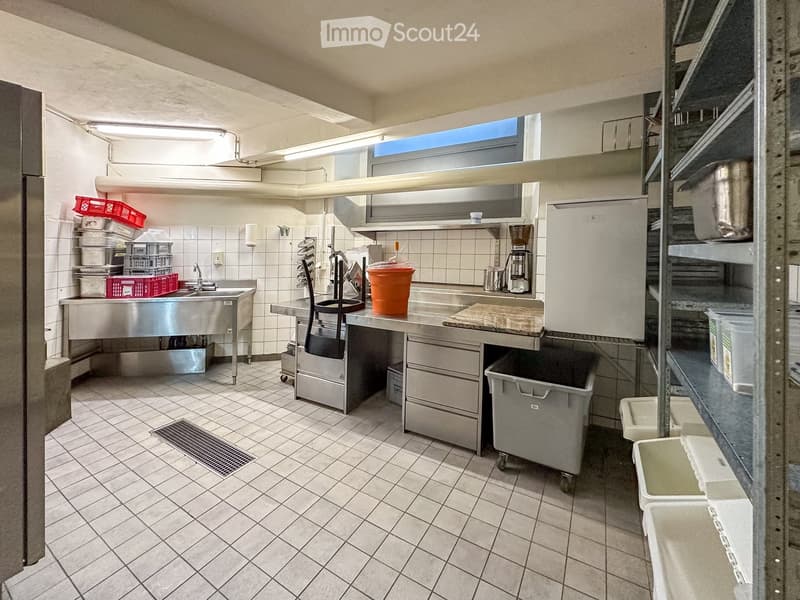Commercial kitchen with stainless steel countertops, sink, refrigerator, coffee machine, storage shelves, and a drain in the floor.