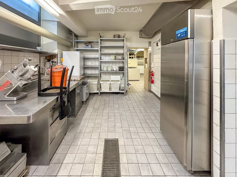 Commercial kitchen with stainless steel appliances, tiled floor, storage shelves, and a refrigerator.