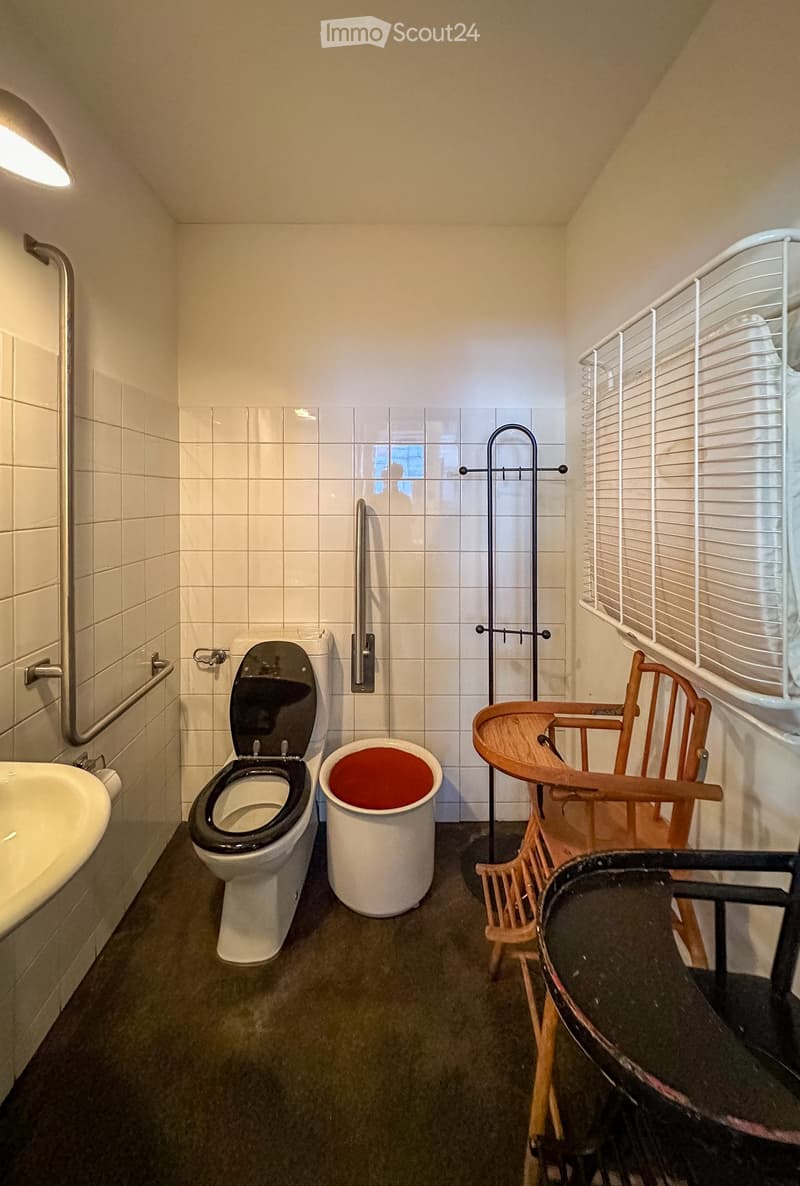 A small bathroom with a toilet, sink, and a bucket. There is a wooden chair and a metal stand in the room.