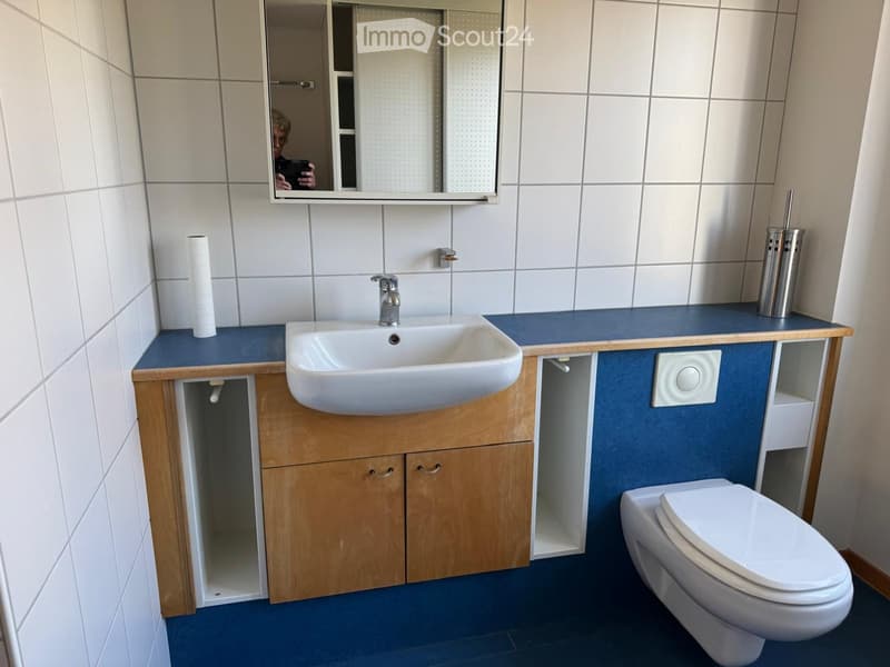 Bathroom with white sink, mirror, blue counter top, white toilet, wall mounted soap dispenser, toilet paper holder, wooden cabinet, and white tiles